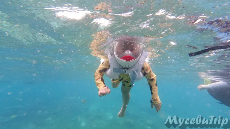 A happy child swimming in the clear blue waters of Hilutungan Marine Sanctuary wearing a shark mask.